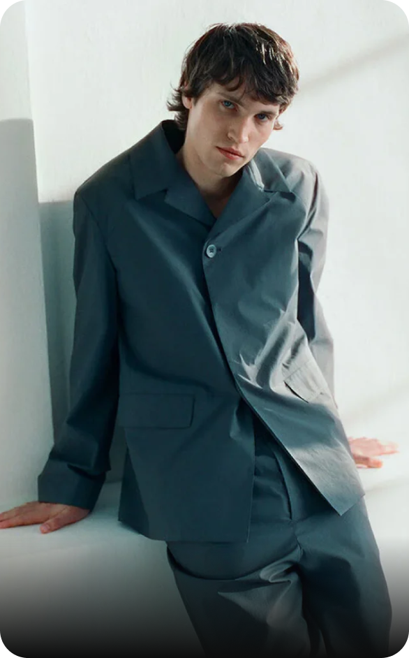 Modelo masculino con traje gris, apoyado en una pared blanca, mostrando estilo moderno y minimalista.