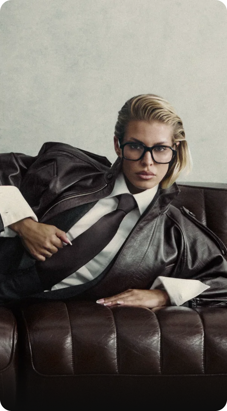 Mujer con gafas y traje de cuero, recostada en un sofá, mostrando un estilo moderno y profesional.