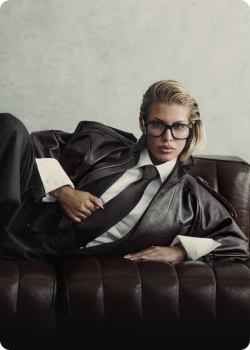 Mujer con gafas y traje negro recostada en un sofá, representando un estilo moderno y elegante.
