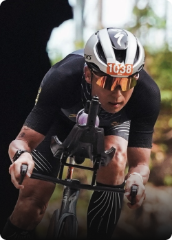 Ciclista en competición, usando casco y gafas, pedaleando rápidamente en un entorno natural.