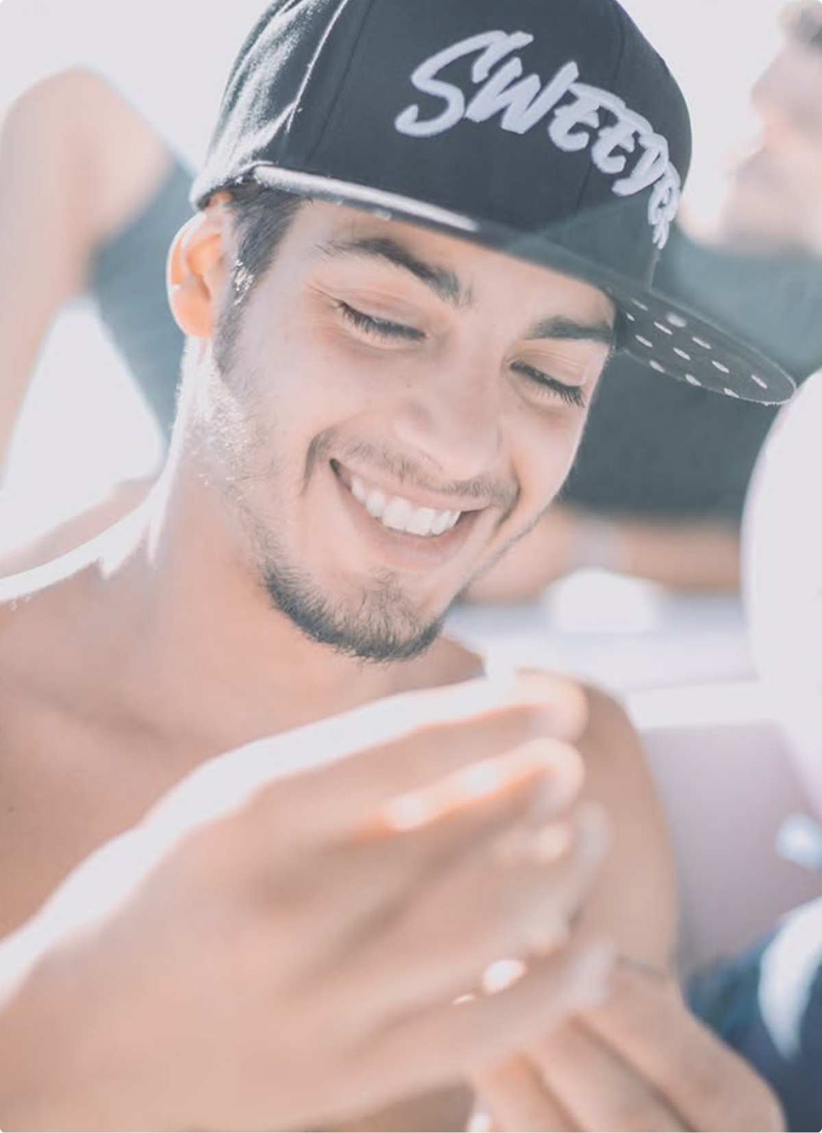 Joven sonriente con gorra negra, observando un objeto en sus manos, en un ambiente relajado.