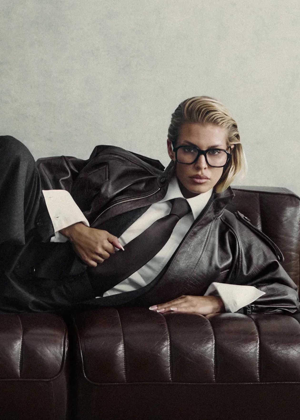 Mujer con gafas y traje oscuro recostada en un sofá marrón, mostrando estilo y elegancia.