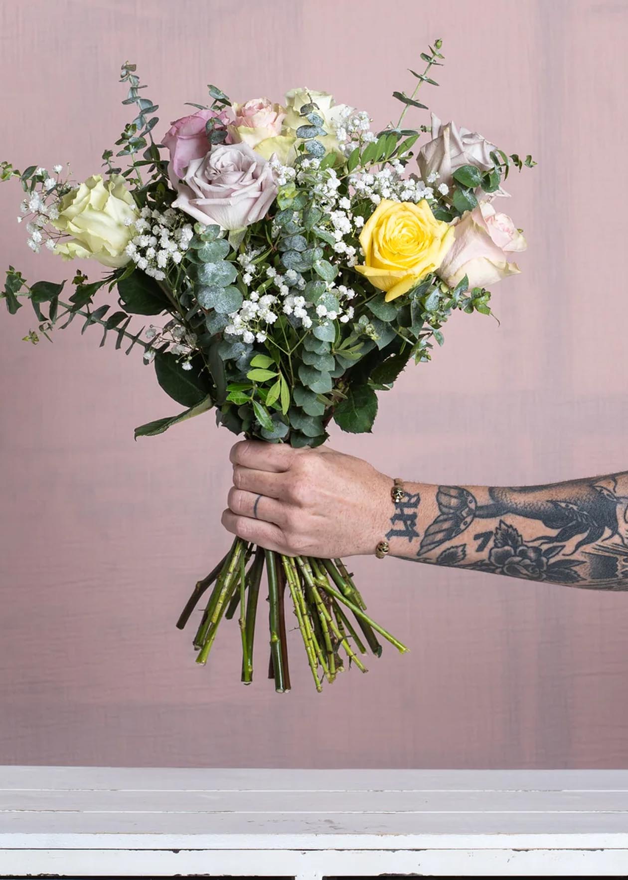 Ramo de flores con rosas en tonos suaves y eucalipto, sostenido por una mano tatuada sobre fondo rosa.
