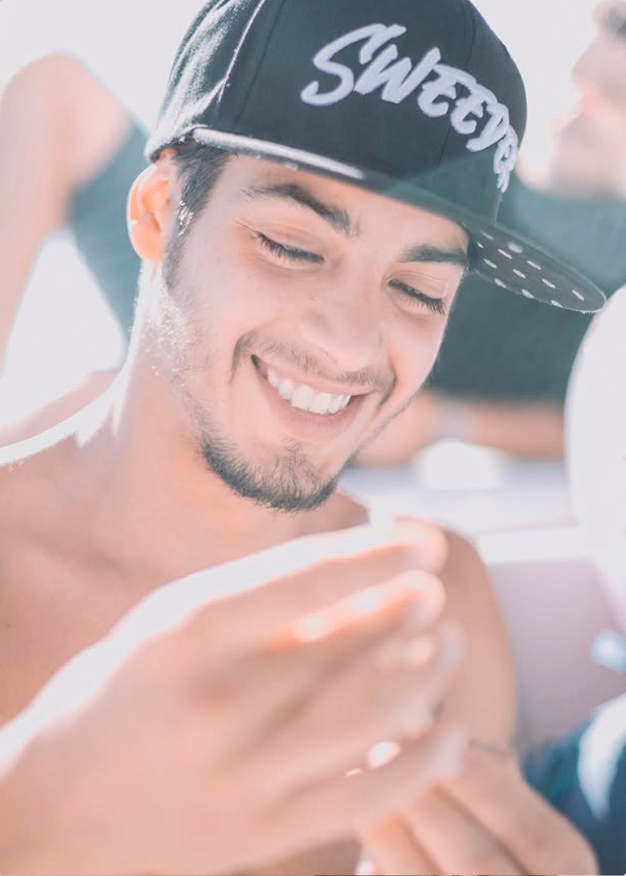 Joven sonriendo mientras sostiene un objeto, con una gorra negra que dice 'Sweeter'.