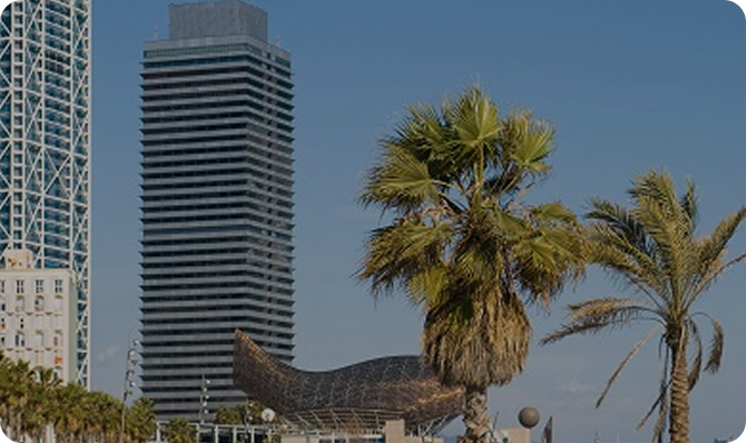 Vista de edificios en Barcelona, incluyendo un rascacielos y una escultura junto a palmeras.