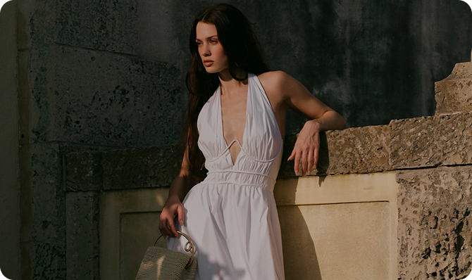 Mujer con vestido blanco posando junto a una pared de piedra, sosteniendo un bolso en su mano.